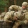Two soldiers in MultiCam/OCP camouflage uniforms are engaged in what appears to be a military obstacle course or training exercise. One soldier (name tape reads 'JONES') is assisting or pushing a second soldier over a wooden wall obstacle. A rifle is visible resting on the wooden barrier, and the setting is an outdoor wooded training area.