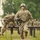 U.S. Army soldiers in full combat gear are navigating an outdoor obstacle course, likely during a training exercise. The foreground soldier, identified by a name tape reading 'REX,' is running through the course wearing OCP (Operational Camouflage Pattern) uniform, body armor, helmet, and carrying a weapon. Additional soldiers are visible in the background completing various obstacles including a wall climb.