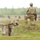 Two soldiers in full combat gear and camouflage uniforms are on a military training range. One soldier is crouching behind a low concrete and brick wall structure, while the other stands on elevated ground observing downrange. Multiple target silhouettes are visible in the background across an open field.