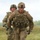 Two U.S. Army soldiers in full combat gear are running across an open grassy field. The lead soldier, identified by a name tape reading 'SPRIGLER,' wears Operational Camouflage Pattern (OCP) uniforms, body armor, helmets, and carries weapons. The image appears to capture a training exercise or competition event.