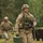 Two U.S. Army soldiers in full combat gear are running across a grassy field, likely during a training exercise. The lead soldier carries a green ammunition can in one hand and an M4-style carbine rifle slung across his body. The background features a wooded tree line and what appear to be wooden obstacle or range markers.