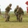 Five soldiers in MultiCam/OCP camouflage uniforms and full tactical gear are gathered on a grassy field, appearing to be engaged in a training exercise or briefing. One soldier is gesturing with a pointed finger, suggesting instruction or direction is being given. A red diamond-shaped range marker with the number '100' is visible in the background.