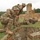 Three soldiers in MultiCam/OCP camouflage uniforms are conducting what appears to be a field training exercise at a military range. Two soldiers are crouched near a deteriorated brick structure while a third is in a prone firing position with a rifle aimed downrange. The setting includes sandy berms, a partially destroyed brick chimney or wall remnant, and a forested background consistent with a European training area.
