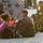 VMFA-115 departs for UDP A group of military personnel in camouflage uniforms are seated on a concrete surface, appearing to rest or wait. A civilian woman in a pink shirt is among the group, suggesting a possible evacuation, humanitarian, or relief operation. The scene appears to take place near military equipment or a vehicle, in warm outdoor lighting.