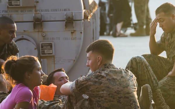 A group of military personnel in camouflage uniforms are seated on a concrete surface, appearing to rest or wait. A civilian woman in a pink shirt is among the group, suggesting a possible evacuation, humanitarian, or relief operation. The scene appears to take place near military equipment or a vehicle, in warm outdoor lighting.