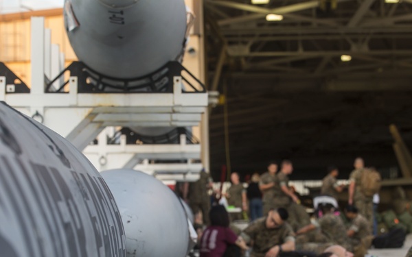 Military personnel in camouflage uniforms are resting on the floor of what appears to be an aircraft hangar or flight line area. Large munitions or ordnance are staged in the foreground on wheeled carts, with partial text visible on the weapons. A mix of military and possibly civilian personnel are gathered in the background near what appears to be a military aircraft.