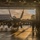 VMFA-115 departs for UDP A military jet aircraft, consistent with an F/A-18 Hornet or Super Hornet, is positioned inside a large aircraft hangar with its hangar doors open at what appears to be sunset or sunrise. The backlighting creates strong silhouettes of the aircraft and surrounding personnel. Multiple ground crew members are visible around the aircraft, suggesting maintenance or pre/post-flight operations.