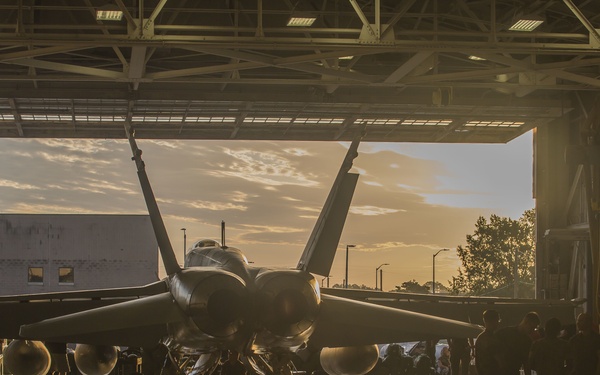 A military jet aircraft, consistent with an F/A-18 Hornet or Super Hornet, is positioned inside a large aircraft hangar with its hangar doors open at what appears to be sunset or sunrise. The backlighting creates strong silhouettes of the aircraft and surrounding personnel. Multiple ground crew members are visible around the aircraft, suggesting maintenance or pre/post-flight operations.