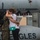 VMFA-115 departs for UDP A person in U.S. Marine Corps camouflage uniform embraces a civilian woman near a military aircraft on a flight line. The emotional embrace, combined with the military context and the service member's backpack, suggests a deployment farewell or homecoming. Partial text on the aircraft reads 'ALTUS' and 'EAGLES'.