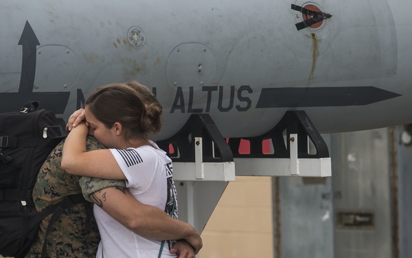 A person in U.S. Marine Corps camouflage uniform embraces a civilian woman near a military aircraft on a flight line. The emotional embrace, combined with the military context and the service member's backpack, suggests a deployment farewell or homecoming. Partial text on the aircraft reads 'ALTUS' and 'EAGLES'.