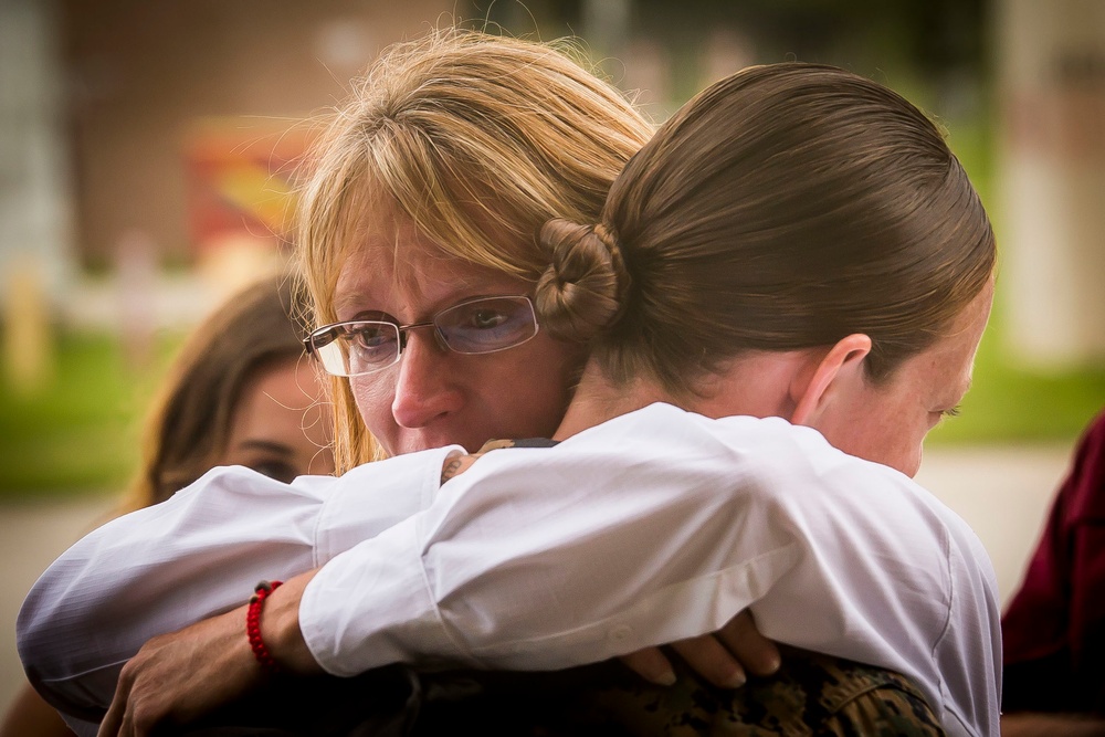 Two individuals are sharing a close embrace in an outdoor setting. The woman facing the camera has blonde hair, glasses, and an emotional expression, suggesting the hug carries significant meaning. A third person is partially visible in the background, slightly out of focus.