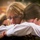 VMFA-115 departs for UDP Two individuals are sharing a close embrace in an outdoor setting. The woman facing the camera has blonde hair, glasses, and an emotional expression, suggesting the hug carries significant meaning. A third person is partially visible in the background, slightly out of focus.