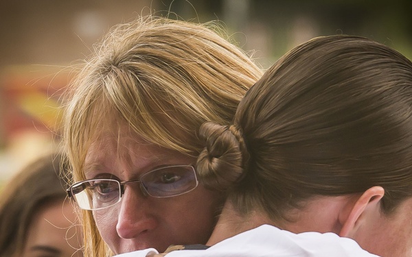 Two individuals are sharing a close embrace in an outdoor setting. The woman facing the camera has blonde hair, glasses, and an emotional expression, suggesting the hug carries significant meaning. A third person is partially visible in the background, slightly out of focus.
