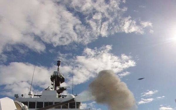 A wide-angle photograph taken from the deck of a military vessel, capturing the moment a deck gun fires, with visible smoke and a projectile in flight. The image shows the ship's superstructure, radar/sensor mast, and various deck equipment under a partly cloudy sky with bright sunlight. The fisheye lens perspective provides a broad view of the ship's forward deck area.