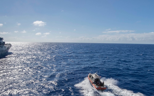 The image shows a maritime operation with a small orange rigid-hull inflatable boat (RHIB) underway at speed on open ocean, viewed from the deck or ramp of a larger vessel. A gray naval warship, likely a U.S. Navy destroyer or frigate, is visible on the left side of the frame. The photo appears to be taken from a ship's well deck or stern ramp area, indicated by the metal plating with rivets visible in the foreground.