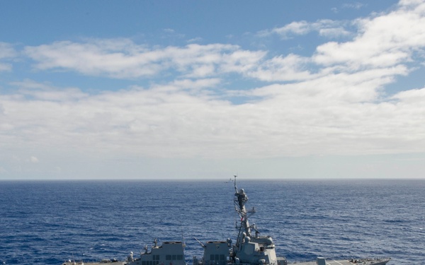 An aerial photograph shows a large U.S. Navy Arleigh Burke-class guided-missile destroyer underway in open ocean under partly cloudy skies. A small rigid-hull inflatable boat (RHIB) or patrol craft is visible alongside the destroyer, moving in the same direction. The ship is painted in standard naval haze gray and appears to be conducting routine operations at sea.