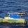 A small green and yellow wooden boat named '2 AMIGOS' with registration number VC5445F is adrift in open blue ocean water. The vessel appears to be in distress or abandoned, with improvised shelter made from a blue tarp and wooden boards visible on deck. The word 'OK' is painted in orange on both the hull and the tarp, likely as a distress signal or status indicator.