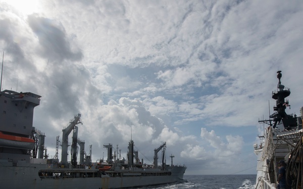 The image shows a replenishment-at-sea (RAS) operation with at least two large naval vessels sailing in close parallel formation on open ocean. The view is taken from the deck of one ship looking toward a large auxiliary/supply vessel on the left, with additional ships visible in the background. The scene depicts a standard underway replenishment evolution conducted by naval forces.