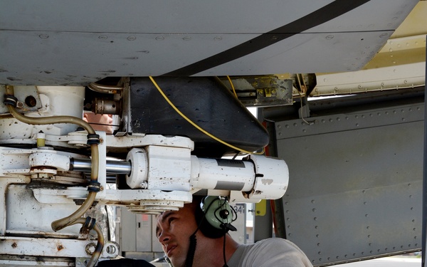 A military aircraft maintainer in US Air Force ABU (Airman Battle Uniform) camouflage is performing maintenance on an aircraft's landing gear assembly. The technician is kneeling beside a large aircraft tire while inspecting or working on the landing gear strut and associated hydraulic components. He is wearing hearing protection headset and black work gloves.