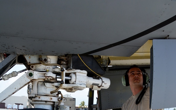 A military aircraft maintainer in US Air Force ABU (Airman Battle Uniform) camouflage is inspecting the landing gear assembly of a large military aircraft on a flight line. The technician is crouching and looking upward at the landing gear strut and mechanical components. The setting appears to be a tropical or warm-climate air base, with palm trees visible in the background.