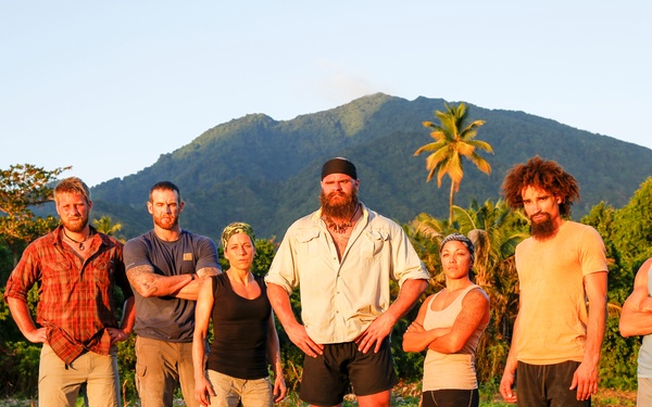 A group of seven people stand together in a rugged outdoor setting with a large mountain and tropical vegetation in the background. They appear weathered and worn, dressed in casual survival-style clothing, posing in a deliberate group formation. The warm golden light suggests either sunrise or sunset.