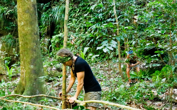 Two individuals are working in a dense tropical forest, constructing or assembling a structure using bamboo or wooden poles lashed together with rope or vines. The foreground person, wearing a black t-shirt, khaki shorts, and a green headband, is actively tying or securing poles at a junction point. A second person is visible in the background, similarly engaged in the construction activity.