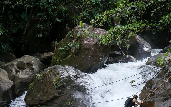 A person is climbing across large boulders beside a rushing waterfall in a lush, tropical environment, using a rope for support and safety. The climber is equipped with a small backpack and is navigating a rocky gorge with white-water rapids below. The scene depicts an adventurous outdoor activity in what appears to be a jungle or rainforest setting.