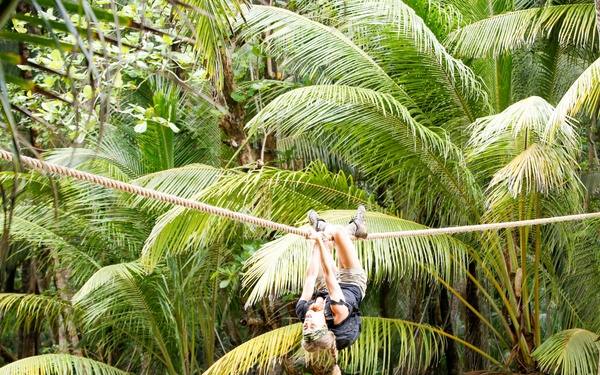 A person is hanging upside down from a rope stretched across a muddy river in a tropical jungle setting. The individual appears to be traversing the rope hand-over-hand while suspended inverted over the water. The lush palm-filled environment suggests a rainforest or jungle adventure activity.