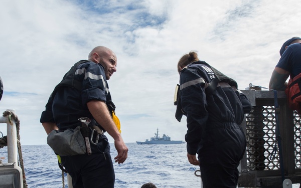 Naval or coast guard personnel are conducting operations on the deck of a vessel at sea. Several crew members in dark uniforms with reflective striping are gathered around an open hatch or boarding ladder, with one person emerging from below or from the water. A second warship is visible on the horizon in the background.