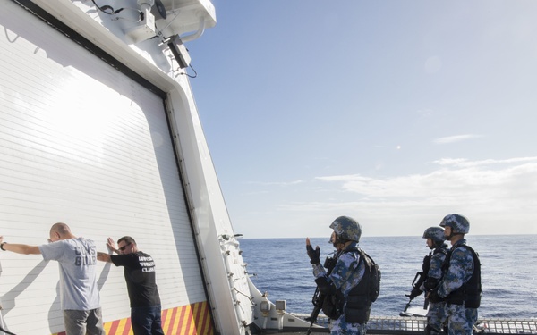 The image shows what appears to be a maritime security or boarding exercise on the deck of a naval or coast guard vessel at sea. Two armed personnel in blue camouflage uniforms and helmets are directing two civilians who have their hands raised against a white bulkhead. The scene takes place on an open ship deck with calm ocean visible in the background.