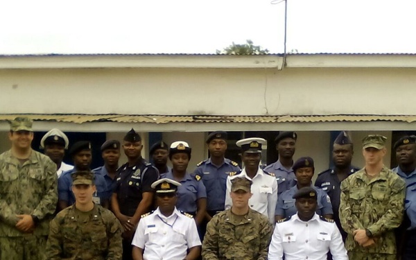 A formal military group photograph featuring personnel from multiple nations in various uniforms, posed in front of a single-story white building. The group includes individuals in US Marine Corps camouflage, US Navy uniforms, and what appear to be West African military/police uniforms in light blue and dark navy. Senior officers are seated in the front row while junior personnel stand in rows behind them.