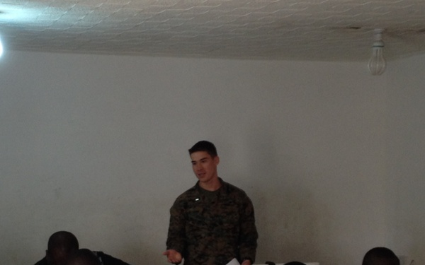 A person in US Marine Corps digital camouflage uniform stands at the front of a computer lab classroom, appearing to instruct a group of students wearing military/naval uniforms. The students are seated at workstations equipped with desktop computers, monitors, and headsets. The setting appears to be a military training facility, likely in an African country based on the visible uniforms and context.