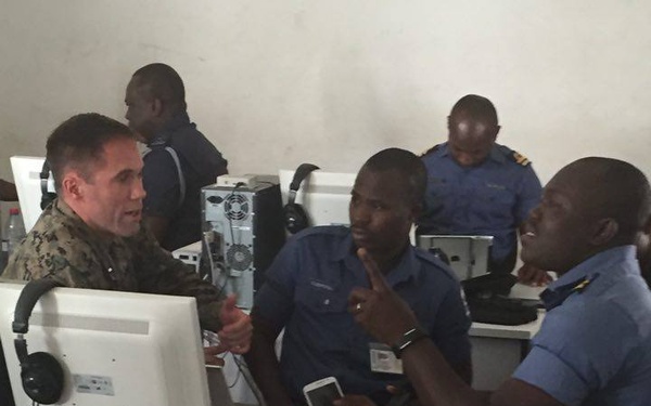 A group of military and uniformed personnel are gathered around computer workstations in what appears to be a training or operations room. One individual in camouflage uniform, likely a Western military advisor, is engaged in discussion with several personnel wearing light blue uniforms, suggesting a joint training or advisory session. The setting appears to be a computer lab or command center environment.