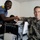 Crisis Response Marines conduct Intelligence training with Ghanaian Forces Two military personnel — one in a blue uniform with shoulder insignia and one in US Marines camouflage — are shaking hands across a desk with computer equipment. A third person is partially visible in the background, leaning over the desk. The setting appears to be an office or training room with white walls.