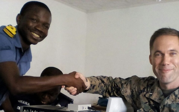 Two military personnel — one in a blue uniform with shoulder insignia and one in US Marines camouflage — are shaking hands across a desk with computer equipment. A third person is partially visible in the background, leaning over the desk. The setting appears to be an office or training room with white walls.