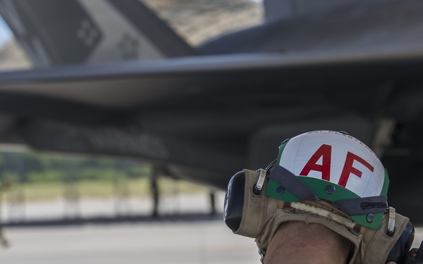 A military ground crew member is positioned beneath a military aircraft on a flight line or tarmac. The individual is wearing hearing protection and a helmet marked 'AF' (Air Force) while holding a handheld radio communicator. Another person in uniform is visible in the background on the tarmac.