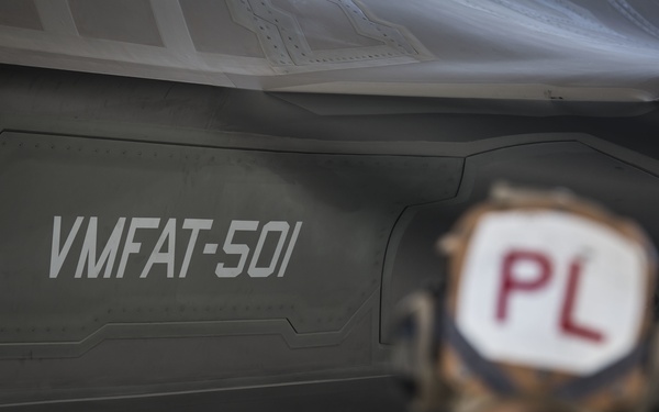 A military personnel member wearing a helmet marked 'PL' stands beneath a stealth aircraft bearing the unit designation 'VMFAT-501' on its fuselage. The image is a close-up shot taken on what appears to be a flight line or tarmac, emphasizing the aircraft's low-observable angular surfaces. VMFAT-501 is a U.S. Marine Corps F-35B training squadron based at MCAS Beaufort, South Carolina.
