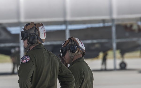 Two military pilots in olive green flight suits and brown flight helmets with oxygen masks stand on an airfield tarmac, appearing to converse. One pilot displays a patch combining the American and British flags on their left shoulder, suggesting a joint US-UK military operation. In the background, what appears to be an F-35 stealth fighter jet is visible along with additional ground personnel.