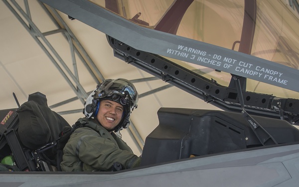A smiling pilot in flight gear sits in the open cockpit of an F-35 fighter jet, numbered '05', bearing U.S. military insignia. The aircraft displays multiple safety markings including ejection seat warnings and rescue instructions. The photo appears to be taken inside or near a hangar structure.
