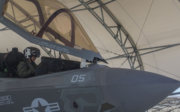 A U.S. military F-35 fighter jet (tail number 05) is shown under a temporary shelter/hangar structure with a pilot seated in the cockpit wearing a flight helmet. Ground crew personnel are visible nearby, with one individual standing to the right in a green flight suit. The aircraft bears U.S. Air Force/Marine Corps roundel markings and a name stencil reading 'CPL KIRKHAM L.E. NORWICH CT' on the fuselage.