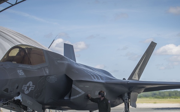 A U.S. military F-35 stealth fighter jet is parked on a flight line apron near a hangar structure. A ground crew member in a flight suit is conducting a pre-flight or post-flight inspection of the aircraft. The setting appears to be a military air base under partly cloudy skies.