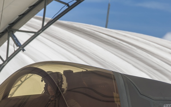 A pilot is seated in the cockpit of an F-35 Lightning II stealth fighter jet, numbered '05', positioned inside or adjacent to a temporary fabric shelter structure. The aircraft's canopy is closed with the pilot visible through the tinted gold-hued canopy glass. Various safety and rescue markings are clearly visible on the fuselage.