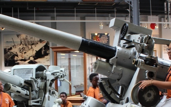 A group of children and a female guide or educator in an orange shirt are visiting a military museum exhibit featuring large anti-aircraft guns or naval artillery pieces. The guide appears to be explaining the weapons to the young visitors, who are wearing matching orange shirts suggesting a school or organized group trip. The exhibit space contains multiple artillery pieces along with historical photographs and display cases.
