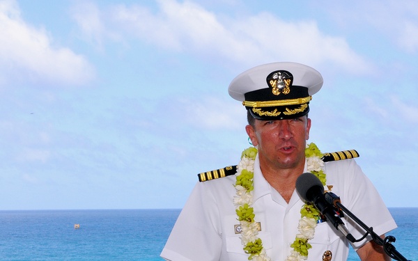 A U.S. Navy officer in white dress uniform is speaking at a podium with a microphone, wearing a traditional Hawaiian lei. The setting appears to be an outdoor ceremony at a coastal location with a turquoise ocean and blue sky visible in the background. The scene suggests a formal military ceremony or event held in Hawaii.