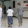 A military service member in Army Combat Uniform (ACU) is assisting an elderly woman using a cane toward a medical area, accompanied by a woman in white scrubs. The setting appears to be a gymnasium or large public building converted into a medical assistance facility. A prominent sign reading 'MEDICAL' with a red arrow pointing left is visible on the wall.