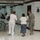 The image shows a medical services area, likely in a school or community center being used as a disaster relief or emergency assistance site. Three individuals are visible in the foreground: a person in white medical scrubs, an elderly woman using a cane, and a soldier in U.S. Army ACU (Army Combat Uniform). Signs on both sides of the hallway read 'Medical Services / Servicios médicos,' indicating bilingual (English/Spanish) services.