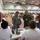 The image shows what appears to be an emergency relief or disaster response operation set up inside a gymnasium or large indoor facility. Two military personnel in camouflage uniforms are interacting with civilian staff seated at a registration or processing table equipped with laptops and a printer. The space is organized with queue barriers and additional seating areas visible in the background.