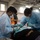 Two medical/dental professionals in blue protective gowns, masks, gloves, and safety glasses are performing a dental procedure on a reclined patient. The setting appears to be a temporary or field medical clinic, likely a community health outreach event, indicated by the large open space with multiple treatment stations visible in the background. A person in a high-visibility safety vest is visible in the background.