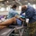 A large-scale dental care event is taking place in what appears to be a convention center or gymnasium. Two dental professionals, one wearing military camouflage pants, are performing a dental procedure on a reclined patient. Multiple other dental stations with providers and patients are visible in the background.