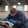 A military dental professional in camouflage uniform and blue medical gown is performing a dental procedure on a patient in what appears to be a field or temporary medical setting. The practitioner is wearing full personal protective equipment including a surgical mask, safety glasses, headlamp, and blue nitrile gloves. An assistant is visible on the left side also wearing gloves and assisting with the procedure.
